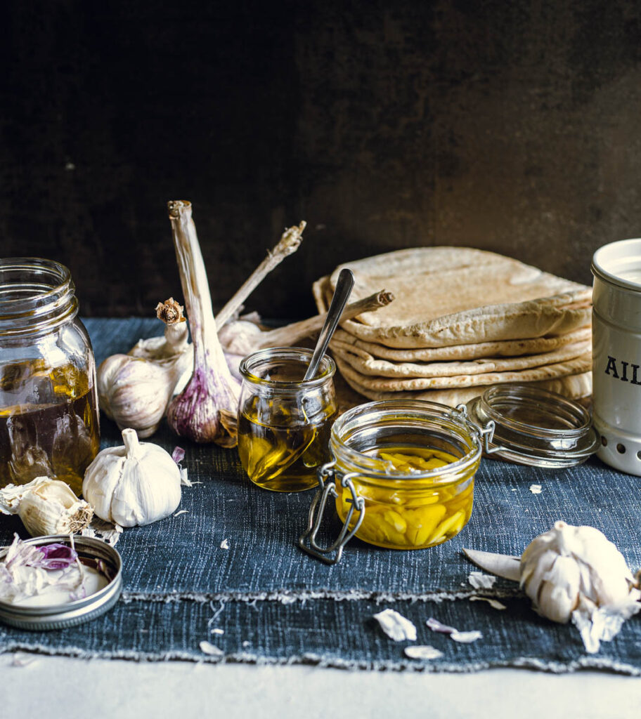 Three small glass jars of garlic-infused oil, surrounded by decorative garlic bulbs, one of which contains garlic cloves cut in half and the others have only the oil