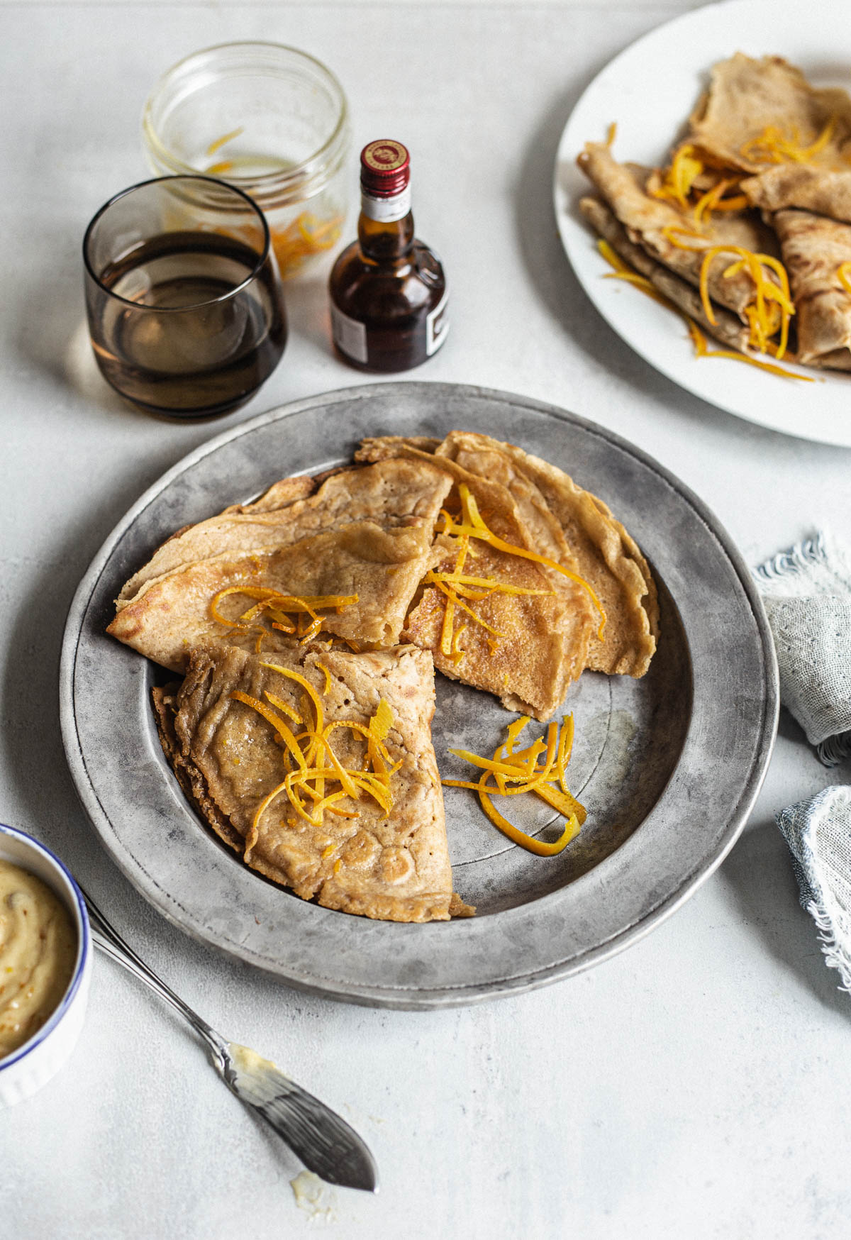 Trois crêpes pliées en quatre et décorées de zeste d'orange dans une assiette en métal accompagnées de beurre moucheté de zeste d'orange dans un ramequin