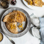 Overhead view of three pancakes folded in four and decorated with orange zest in a metal plate accompanied by butter speckled with orange zest in a ramekin