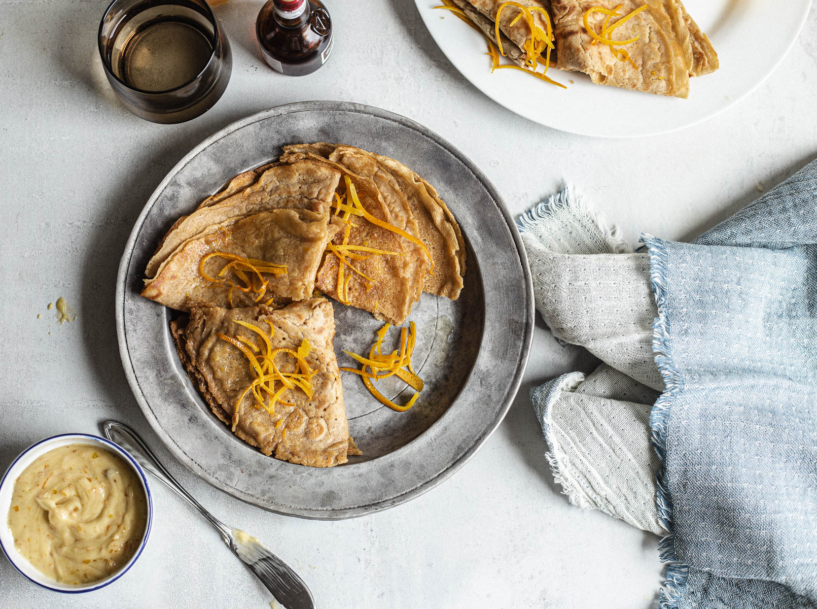 Overhead view of three pancakes folded in four and decorated with orange zest in a metal plate accompanied by butter speckled with orange zest in a ramekin