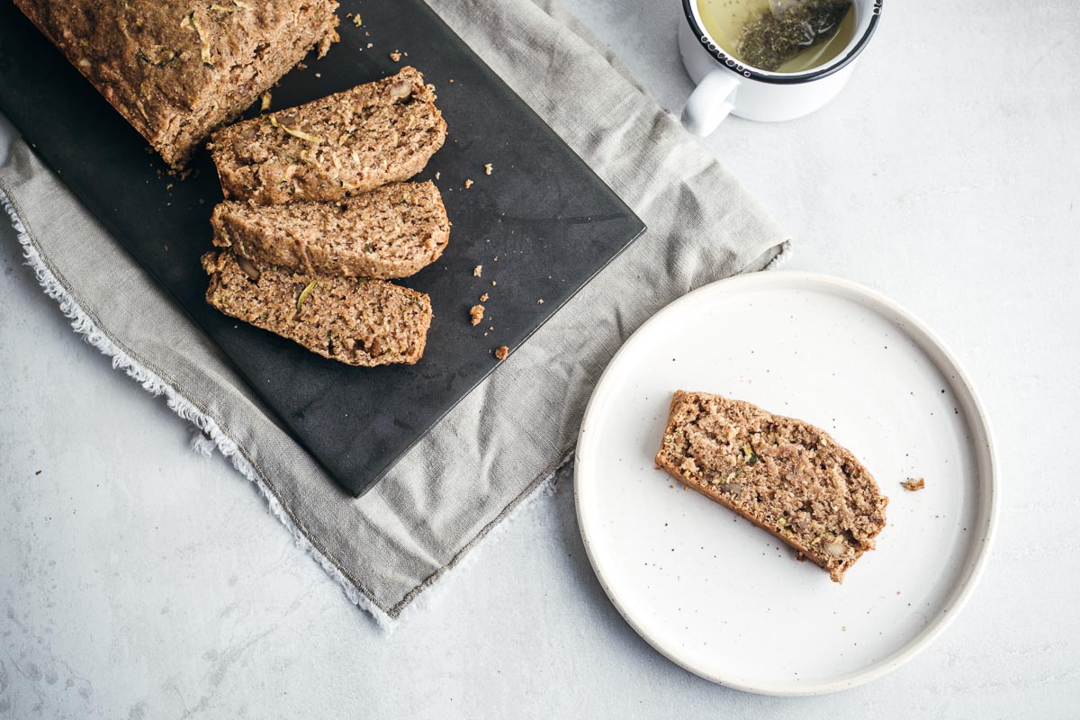 Vegan zucchini bread slice in a dessert dish next to zucchini bread loaf cut in slices