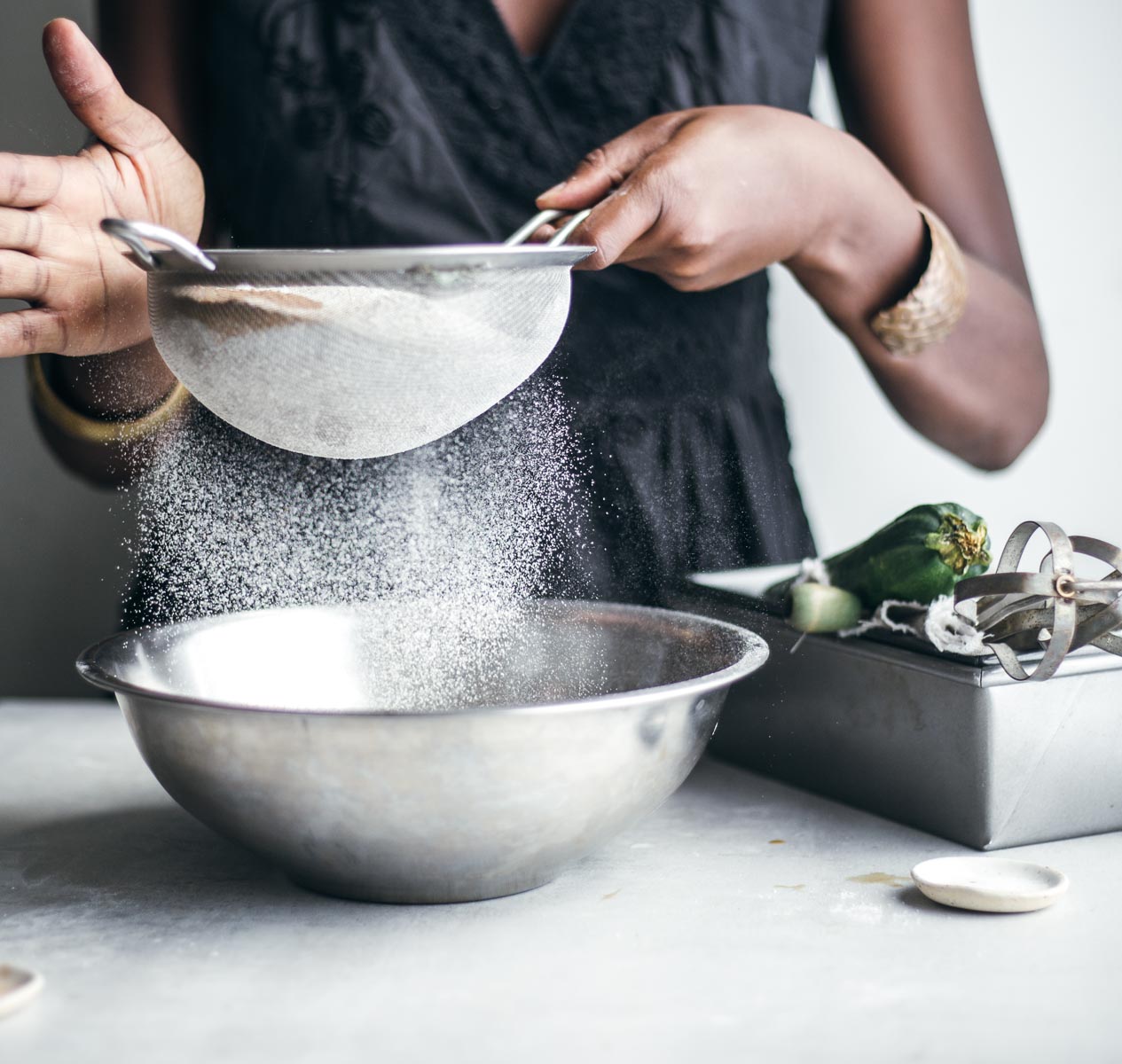 Person in the background filtering the flour through a fine-mesh sieve, dropping it into a mixing bowl