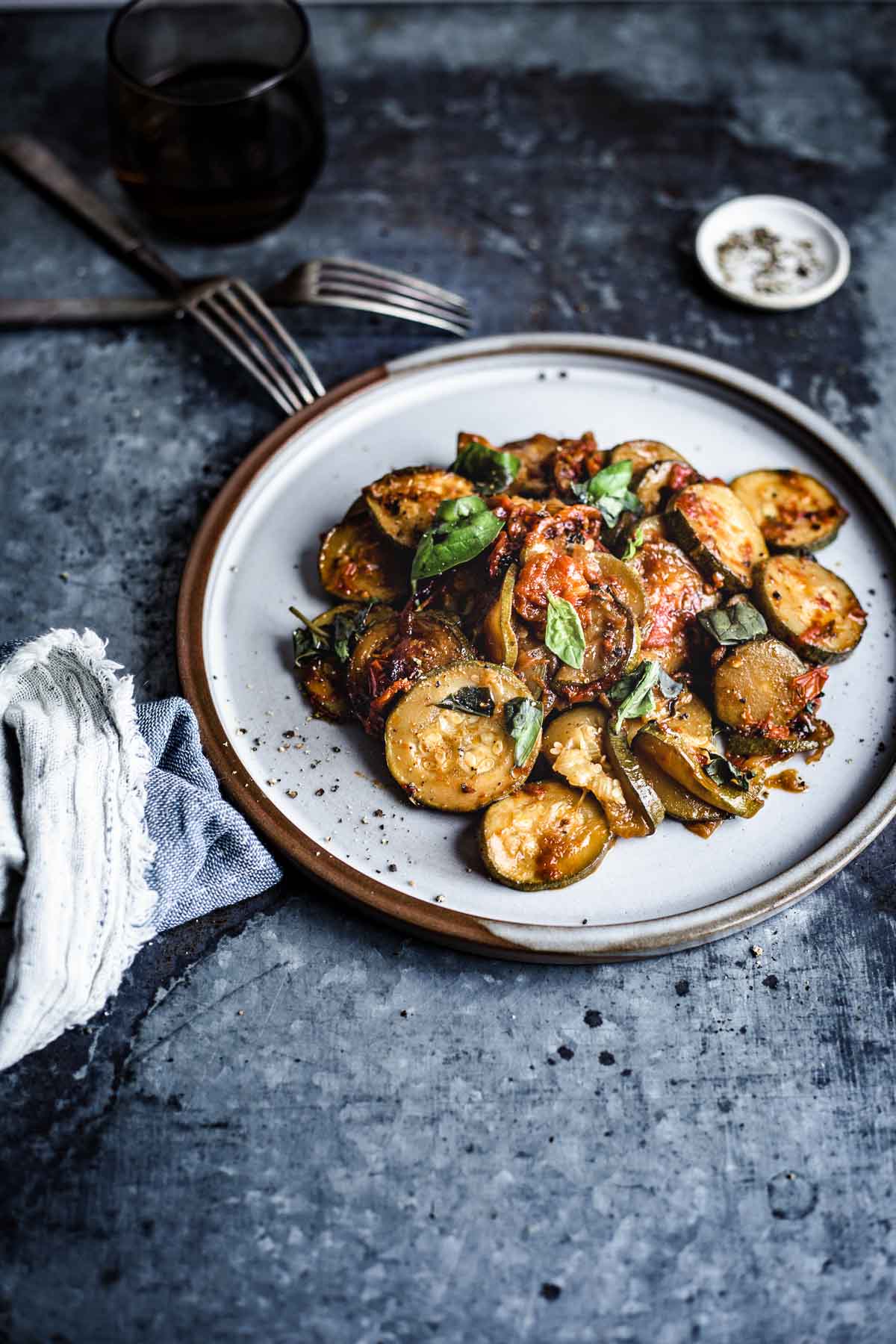 ¾ View of a dish of sliced ​​zucchini, coated in tomato sauce and garnished with torn basil leaf on a round stoneware plate and resting on a metal plate