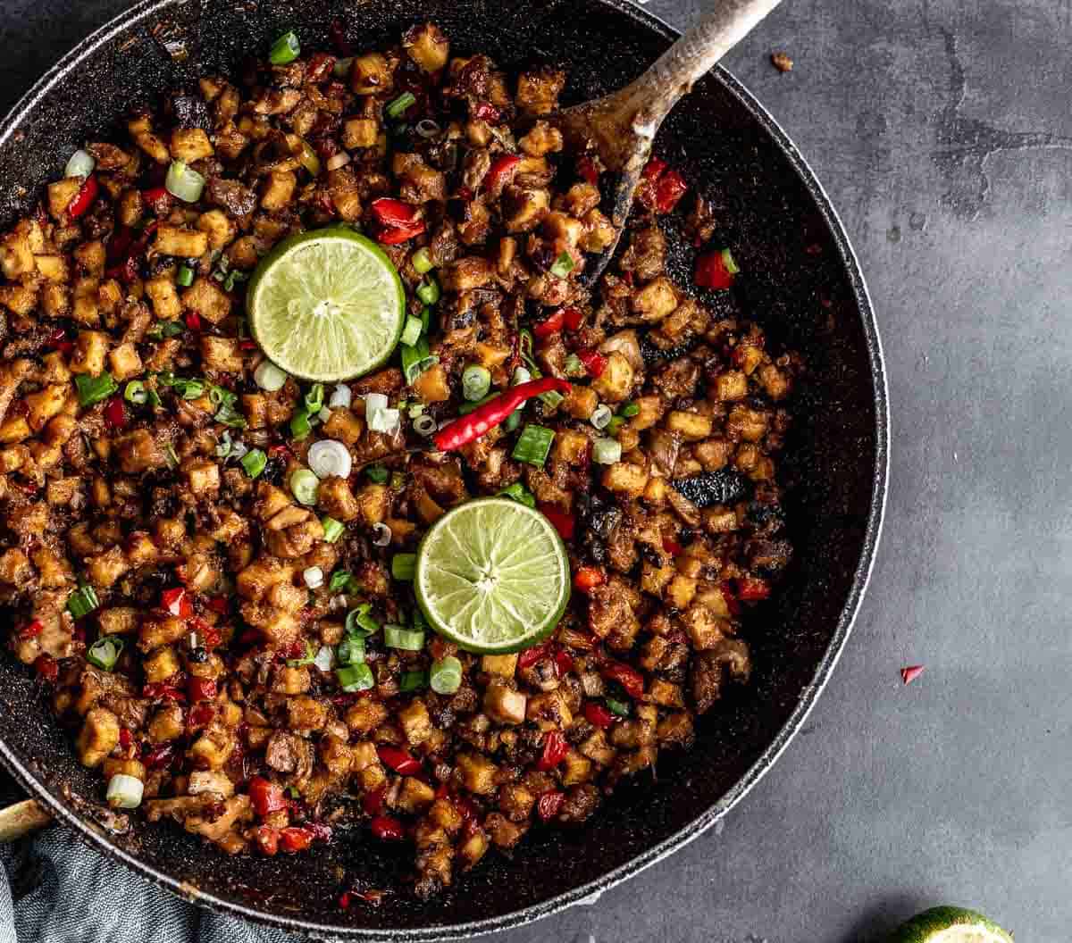 Tofu sisig in a skillet decorated with a halved lemon, chopped green onon and thai chilies.